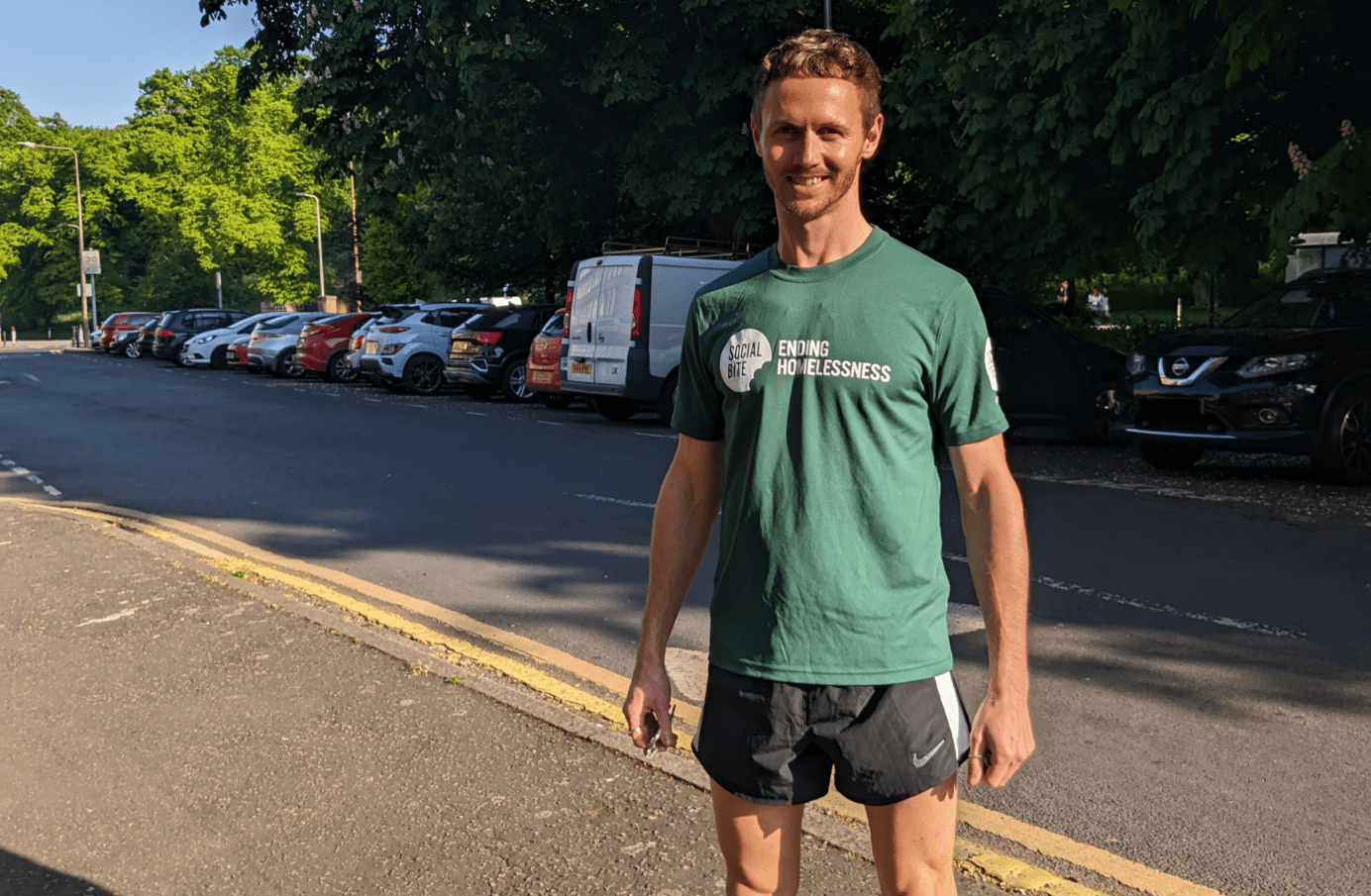 A man in a green tshirt and black running shorts stands on the pavement smiling