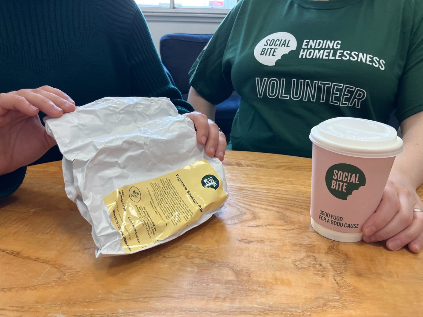 Two people sit together with a coffee. One wears a green tshirt that says volunteer.