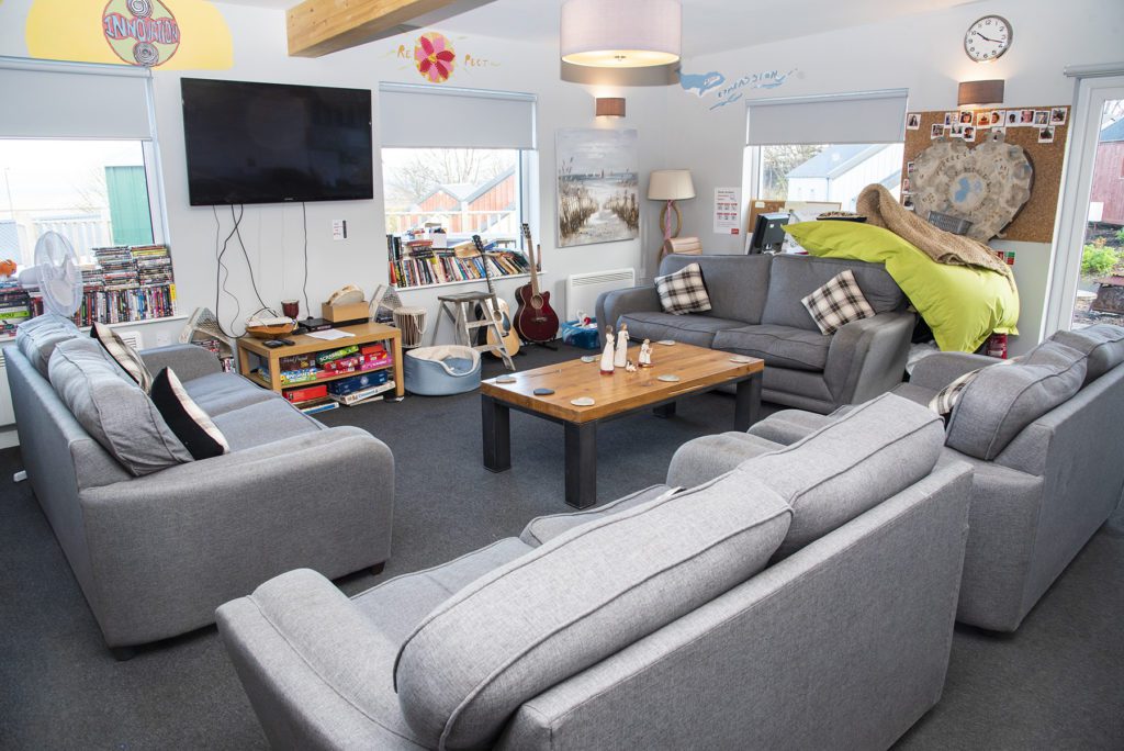 A bright living room with four grey two seater-sofas, a television, books, coffee table, artwork, guitars and three windows looking on to the street.