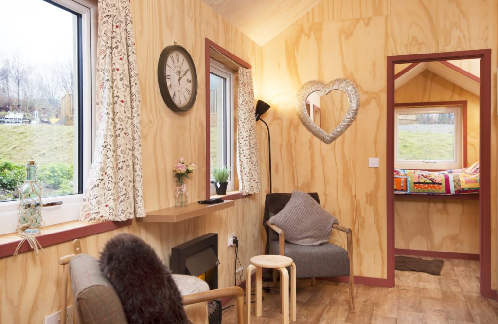 The inside of a wooden cabin with two armchairs either side of a fireplace, a clock on the wall and flowers in vases on the window sill