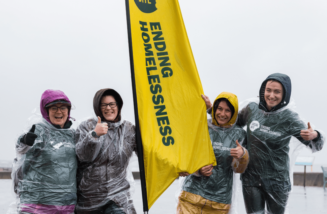 Four women in raincoats and clear ponchos smile and give thumbs up in very wet weather. They have a tall yellow banner between them that reads 'ending homelessness'