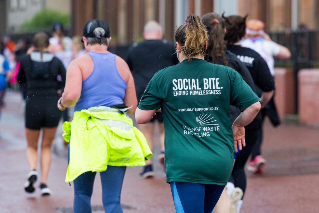 Two women run in event race, away from the camera. One has a dark green tshirt that reads 'Social Bite'