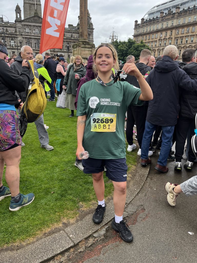 A young woman in a Social Bite tshirt poses with her medal in front of a crowd of people after completing a 10k race