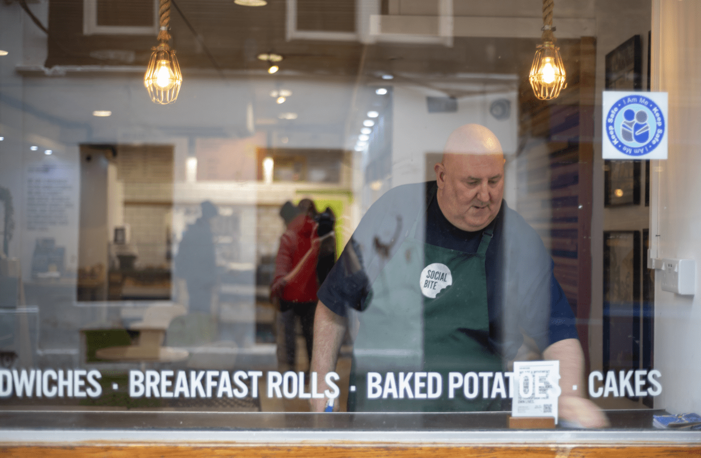 A tall bald man in a green apron tidies a cafe table, viewed from behind a window