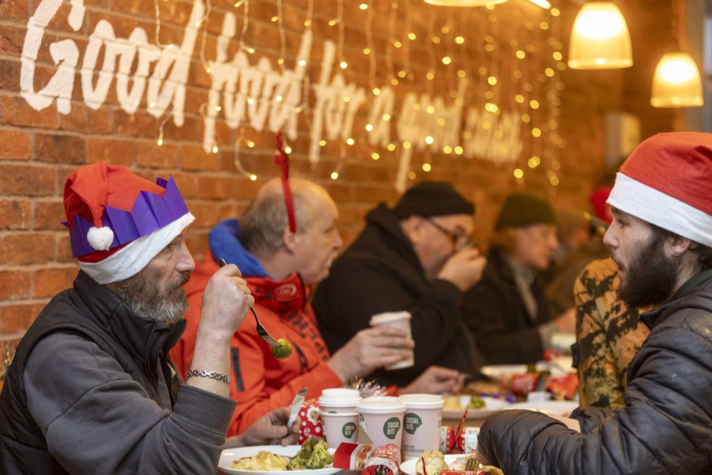 Men in Santa hats and paper crowns eat a meal at a long table, with sparkling lights and Christmas crackers around them